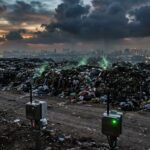 Green methane vapor rises from landfill cracks beside monitoring sensors at dusk.