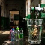 A glass of water beneath a school fountain with corroded pipes visible behind it.