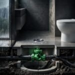Bathroom drain and toilet above pipes with a green backwater valve blocking sewage flow.