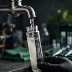 A gloved hand collects water from a kitchen tap into a clear vial.