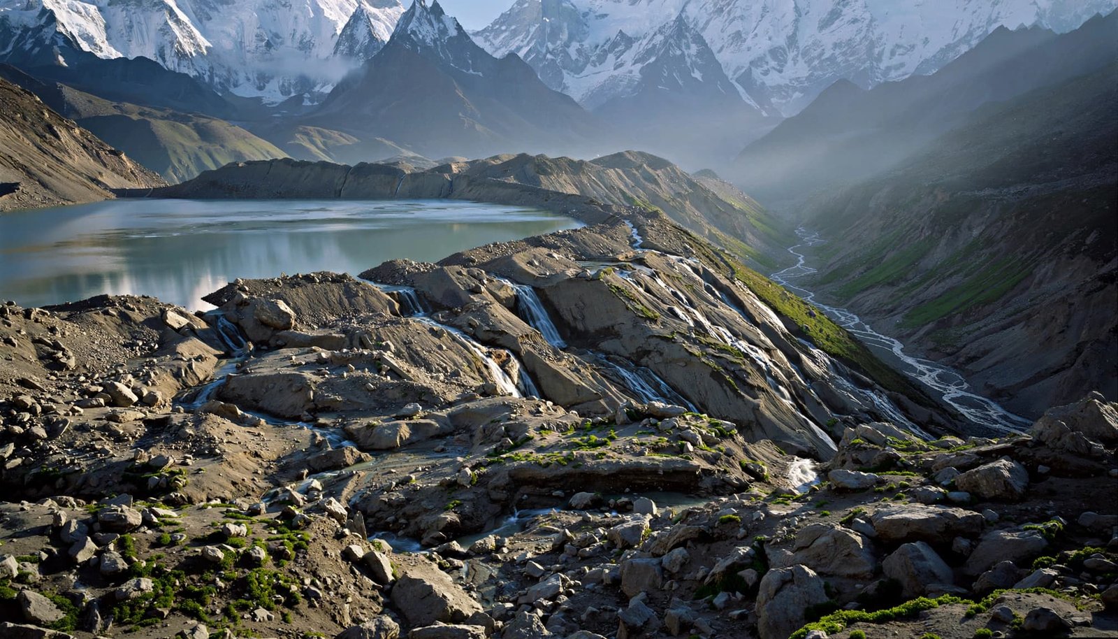 A wide view of a glacial lake held by a moraine dam with waterfalls and snowy peaks.